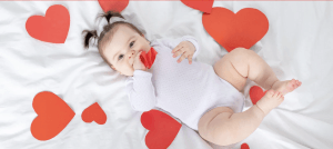 Baby in white onesie laying on a white background surrounded by hearts.